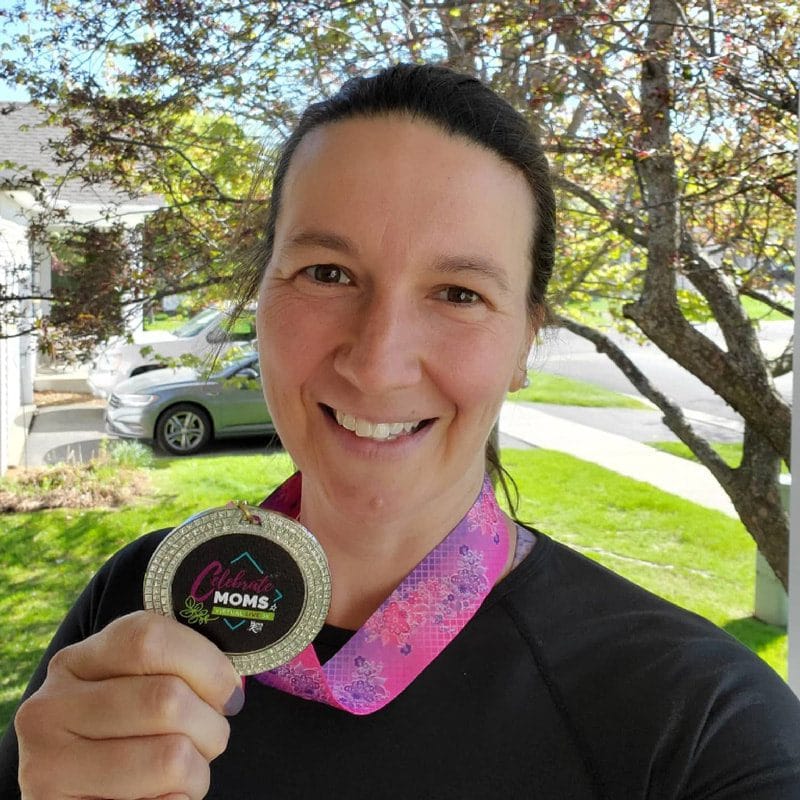 MOTR member in a black shirt on her driveway holding a Celebrate Moms race medal smiling