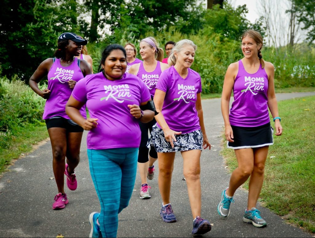 A group of women wearing matching purple Moms on the Run shirts conversating and laughing with jogging on a path