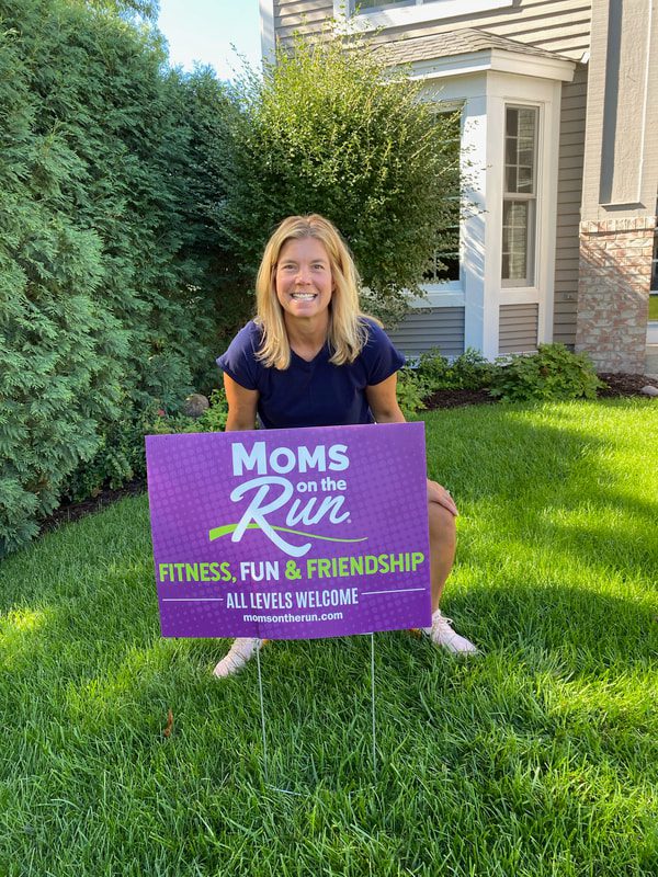 Coach heather, from with Chanhassen and Chaska Moms on the Run, squats behind a Moms on the Run yard sign. These franchises are owned by Katie W.