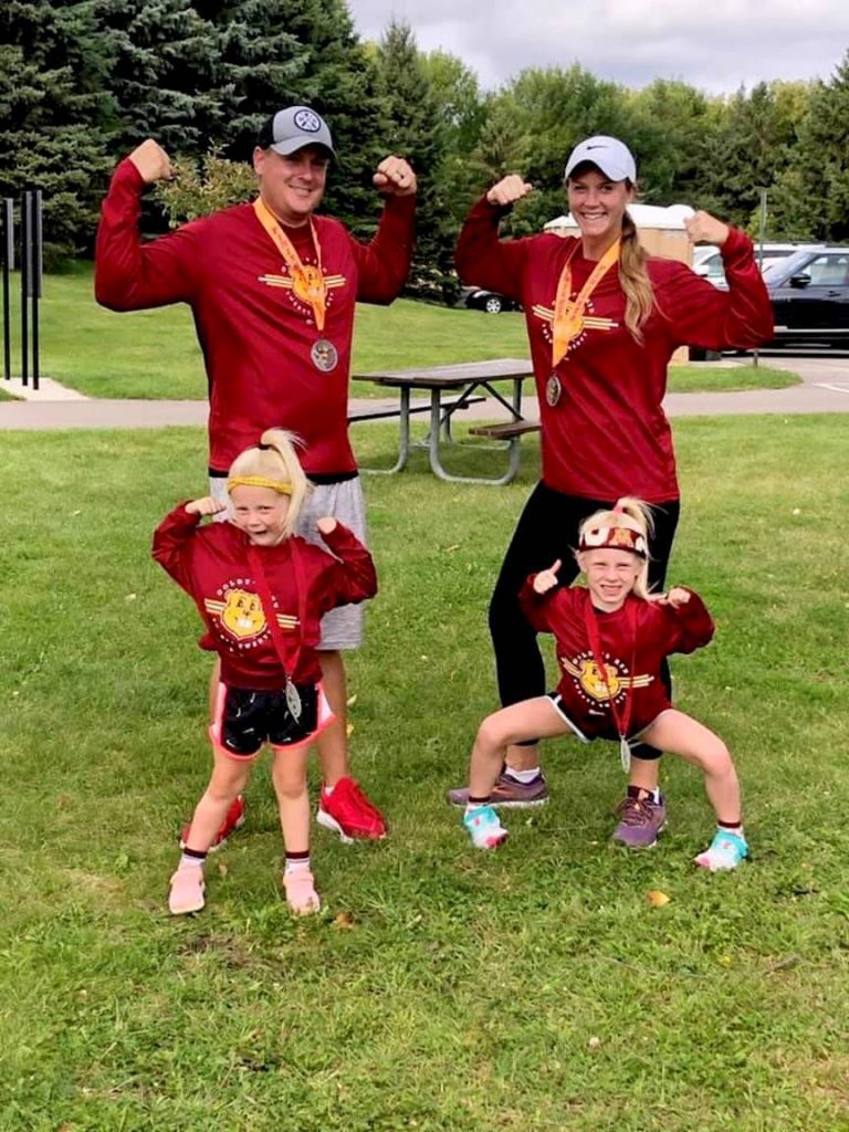 Coach Alicia from Chanhassen and Chaska stands with her husband and two young kids. They are wearing matching shirts and race medals.