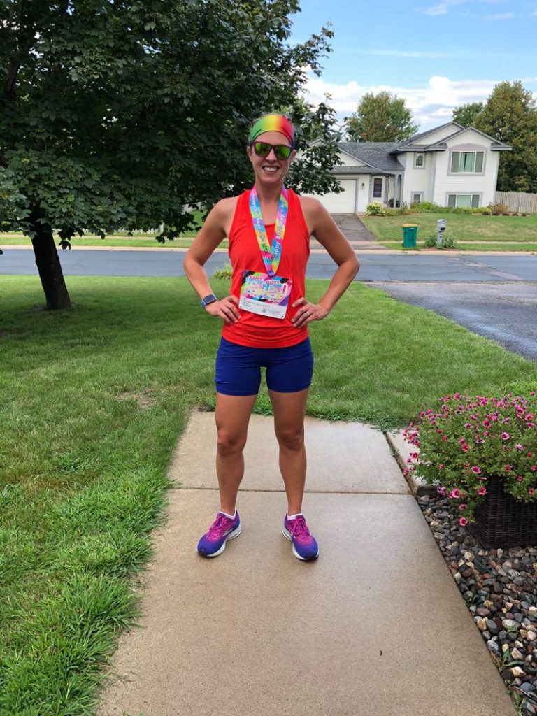 Coach Margaret, from the Chanhassen and Chaska Moms on the Run locations, stands on a sidewalk with her arms triumphantly on her hips. She is wearing a race bib and medal.