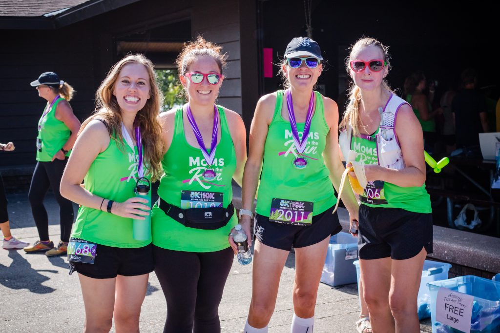 Four Moms on the Run members lined up together and smiling. They're wearing green Moms on the Run shirts, race bibs and race medals.