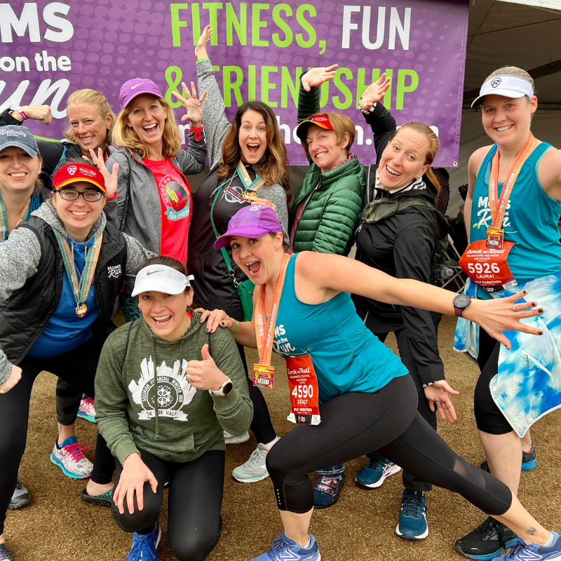 Goup of Moms on the Run members in front of a MOTR banner. They are smiling, posing and wearing race bibs/medals.